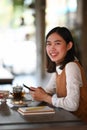 Young female holding mobile phone and smiling to camera while sitting in coffee shop. Royalty Free Stock Photo