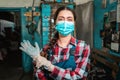 Portrait of a young female mechanic in a uniform and medical mask, wearing work gloves. In the background is a car repair shop Royalty Free Stock Photo