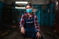 Portrait of a young female pretty mechanic in a work uniform is sitting on a chair, with a medical mask on her face. Indoor.