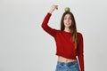 Portrait of young funny fashionable girl in trendy cropped top trying to grab apple placed on her head with chopsticks