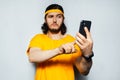 Portrait of young funny guy taking selfie with smartphone on background of grey wall. Man wearing yellow shirt. Royalty Free Stock Photo