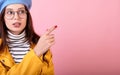 Elegant girl in a stylish beret hat and a yellow cloak in glasses shows a finger to the right on a pink background Royalty Free Stock Photo