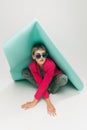 Portrait of young girl in bright jacket and vintage pants posing, sitting under big paper piece isolated over grey Royalty Free Stock Photo