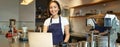 Portrait of young girl cafe owner, looking at her laptop, taking order, serving customer in coffee shop