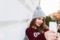 Portrait young girl with long hair in marsala sweater making selfie on grey wall background on street. She wears knitted