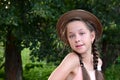 Portrait of young girl with two braids and hat, posing against background of green foliage. Royalty Free Stock Photo