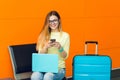 Portrait of young girl in yellow sweater sitting on chair on orange background. She has blue laptop on knees and blue Royalty Free Stock Photo