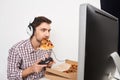 Portrait of young good-looking male gamer playing computer games with controller in headphones, holding pizza in mouth Royalty Free Stock Photo