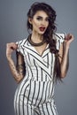 Portrait of young gorgeous dark-haired tattooed lady in striped overall and splendid necklace holding sleeves in her hands