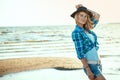 Portrait of young gorgeous blond model in black felt hair, checked blue shirt and denim shorts standing at the seaside
