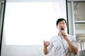 Portrait of young handsome Asian male speaker publicly speaking on stage to group of audience with white board behind.