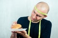 Portrait of a young handsome bald man with a measuring tape eating a high-calorie big burger on a plate with a fork Royalty Free Stock Photo