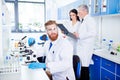 Portrait of young handsome bearded stylish intern, who is wearing gloves and labcoat, safety glasses. Behind him are people who