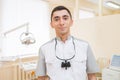 Portrait of Young handsome doctor with dental binocular loupes on at dentist clinic smiling and looking at camera.