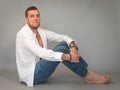 Portrait of a young handsome man dressed in a white unbuttoned shirt and blue jeans sitting on floor in studio on grey Royalty Free Stock Photo