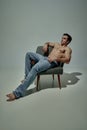 Portrait of young handsome man with muscular relief body sitting on chair, posing shirtless in pants over pale green Royalty Free Stock Photo