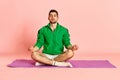 Portrait of young handsome man sitting on sports mat in yoga pose and meditating against pink studio background Royalty Free Stock Photo