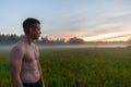 Profile view of young handsome muscular man shirtless against grass field with fog in the break of dawn