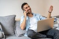 Portrait of young, handsome, smiling man talking on the phone and using his laptop while sitting on the sofa at home. Royalty Free Stock Photo