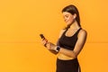 Portrait of young happy attractive sporty woman in black sportwear standing near orange wall background and holding phone, reading Royalty Free Stock Photo