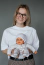 Portrait of young happy beauteous woman holding baby doll in white clothes with two hands on grey background. Studio.