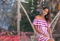 Portrait of a young happy beautiful woman in a striped dress sitting and swinging on a wicker hammock, looking away. Sand in the Royalty Free Stock Photo