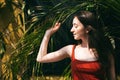 Portrait of a young happy brunette woman in a red t-shirt in profile with a leaf from a palm tree. Face without makeup