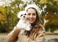 Portrait of young happy Caucasian teenager girl in french white hat and warm coat holding and hugging her little cute white dog Royalty Free Stock Photo