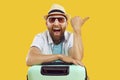 Young man in sunglasses and beach hat with suitcase pointing to the side on yellow background. Royalty Free Stock Photo