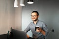 Portrait of young happy man working home at laptop, holding black coffee cup in hand, wearing eyeglasses and shirt, sitting at Royalty Free Stock Photo