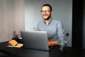 Portrait of young happy man working home at laptop, wearing eyeglasses and shirt, sitting at desk, background of grey wall Royalty Free Stock Photo