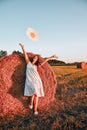 Portrait of young happy woman flying hat on the haystack in morning sunlight, countryside. Beautiful woman in a dress sits on a Royalty Free Stock Photo