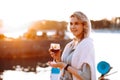 Portrait of young happy woman with long wavy fair hair, standing on embankment, holding glass of red wine at sunset. Royalty Free Stock Photo