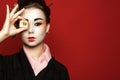 Portrait of young heisha in kimono holding a piece of sushi in front of her eye on red background.