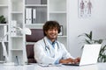 Portrait of young hispanic male medical doctor wearing white coat sitting at desk with laptop in office and smiling at Royalty Free Stock Photo