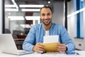 Portrait of a young hispanic man sitting at a desk in a modern office, holding an open letter in an envelope and smiling Royalty Free Stock Photo