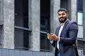 Portrait of a young Indian male businessman smiling at the camera, holding a mobile phone and standing on the street Royalty Free Stock Photo