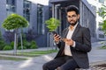Portrait of a young Indian man in a business suit sitting on a bench near an office center, holding a credit card and a Royalty Free Stock Photo