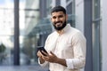 Portrait of a young Indian man in a casual shirt standing near an office center, holding a mobile phone and looking at Royalty Free Stock Photo