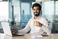 Portrait of a young Indian man sitting at an office desk, working on a laptop, taking a break, holding a cup in his hand Royalty Free Stock Photo