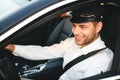 Portrait of young joyful man taxi driver in uniform and cap, driving car wearing seat belt Royalty Free Stock Photo