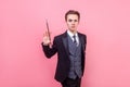 Portrait of young magician in tuxedo standing with raised magic wand. indoor studio shot isolated on pink background