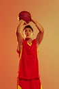 Portrait of young man, concentrated basketball player throwing ball into basket isolated over orange studio background Royalty Free Stock Photo