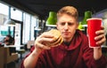 Portrait of a young man eating a burger and drinking a drink at a fast food restaurant. Student holds a glass of drink and an Royalty Free Stock Photo