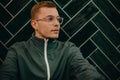 Portrait of young man in jacket and glasses sitting agiant dark tiled wall.