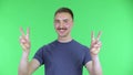 Portrait of a young man shows two fingers victory gestur. Cute male with a mustache in a blue t-shirt posing on a green
