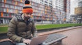 Portrait of young man, sitting on the bench and working at laptop on background of modern city buildings. Wearing orange hat. Royalty Free Stock Photo