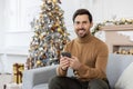 Portrait of a young man sitting at home on the couch near Christmas decorations and a Christmas tree, holding a phone Royalty Free Stock Photo