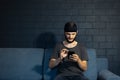 Portrait of young man, sitting on sofa, using screen of smartphone. Background of black brick wall. Royalty Free Stock Photo