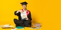 Portrait of young man, student in graduation cap and gown sitting on floor around books isolated over yellow studio Royalty Free Stock Photo